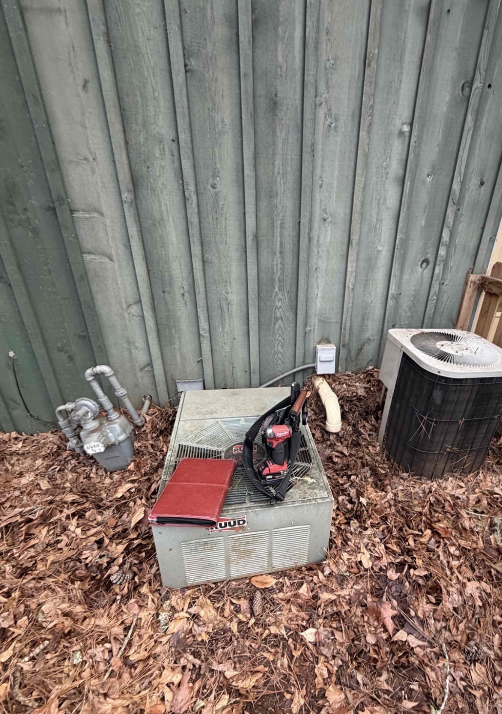 Old Ruud condenser unit completely buried in leaves and debris, sitting directly on the ground with no pad, well past its service life